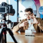 selective focus of blogger in front of digital camera pointing on smartphone at table in coworking